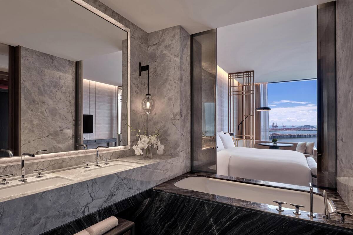 A modern bathroom with a large mirror, dual sinks, a bathtub, and a view of a bedroom with a bed and a cityscape.