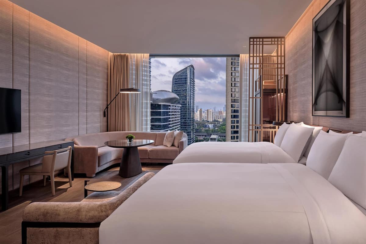 A modern hotel room with a large bed, a sofa, a desk, and a view of the city skyline.