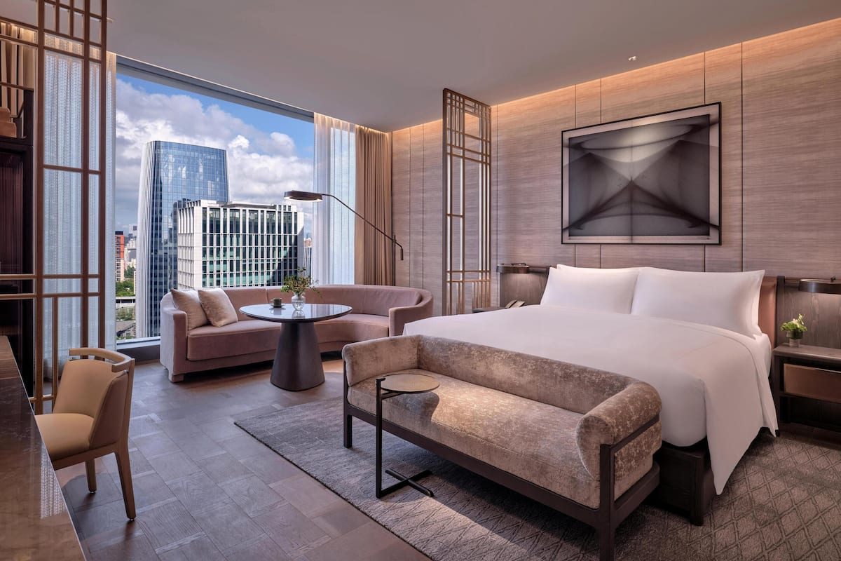 A modern hotel room with a large bed, a sofa, a small table, and a chair. There is a large window with a city view.