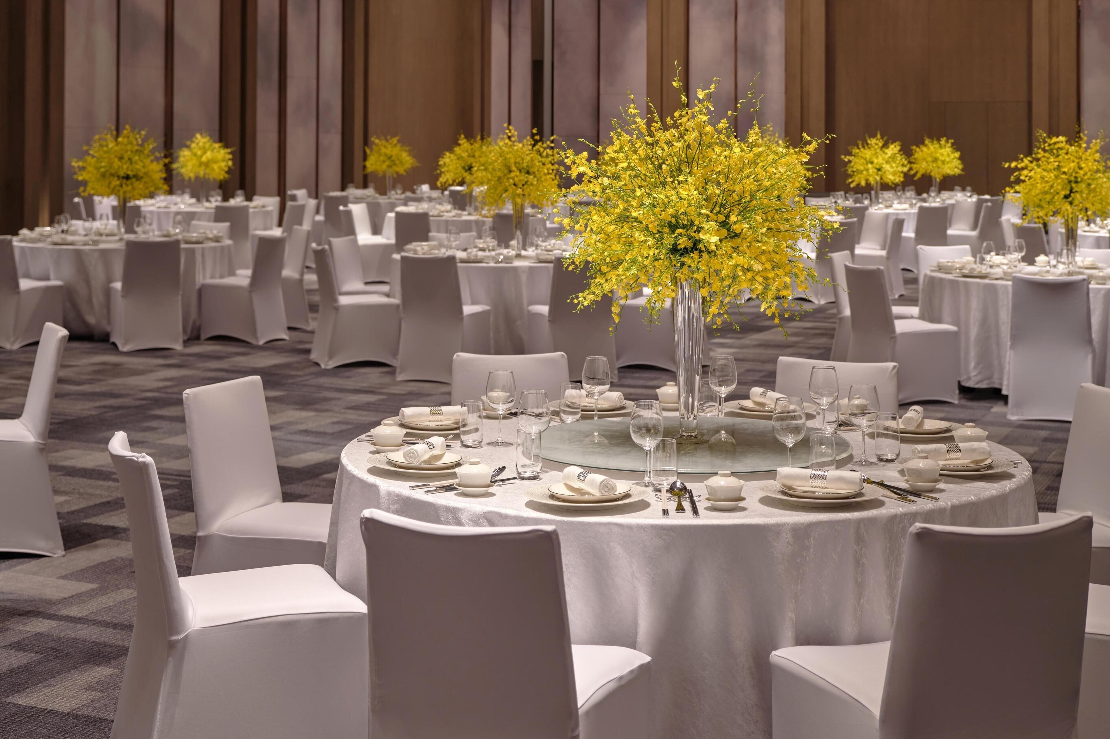 Une salle de banquet avec des tables rondes dressées pour le repas, chacune étant recouverte d’une nappe blanche et entourée de chaises.