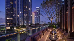 Terraza en la azotea con un sofá y sillas, un árbol y vistas de edificios altos al anochecer.