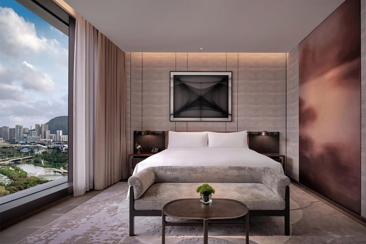 A modern hotel room with a large bed, a view of a cityscape, and a city park.