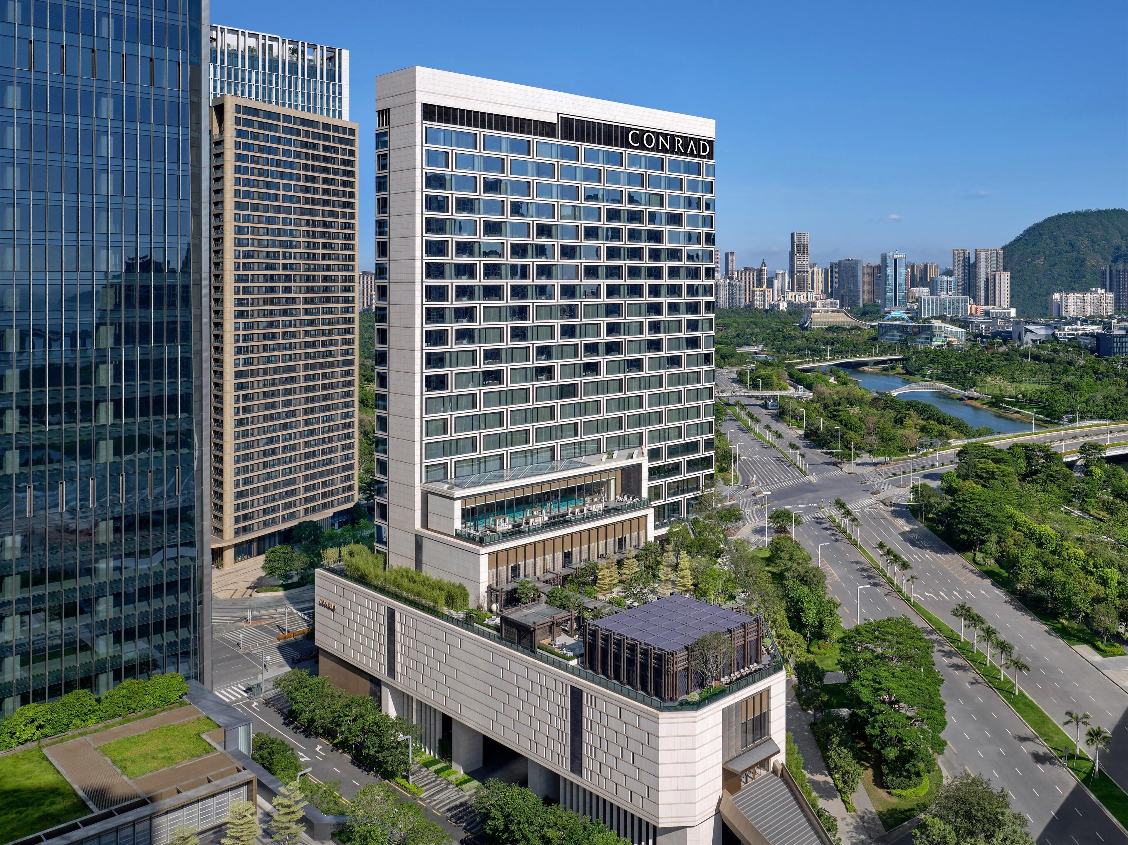 Un edificio moderno de gran altura con un llamativo letrero 'CONRAD', rodeado de exuberante vegetación y un río.