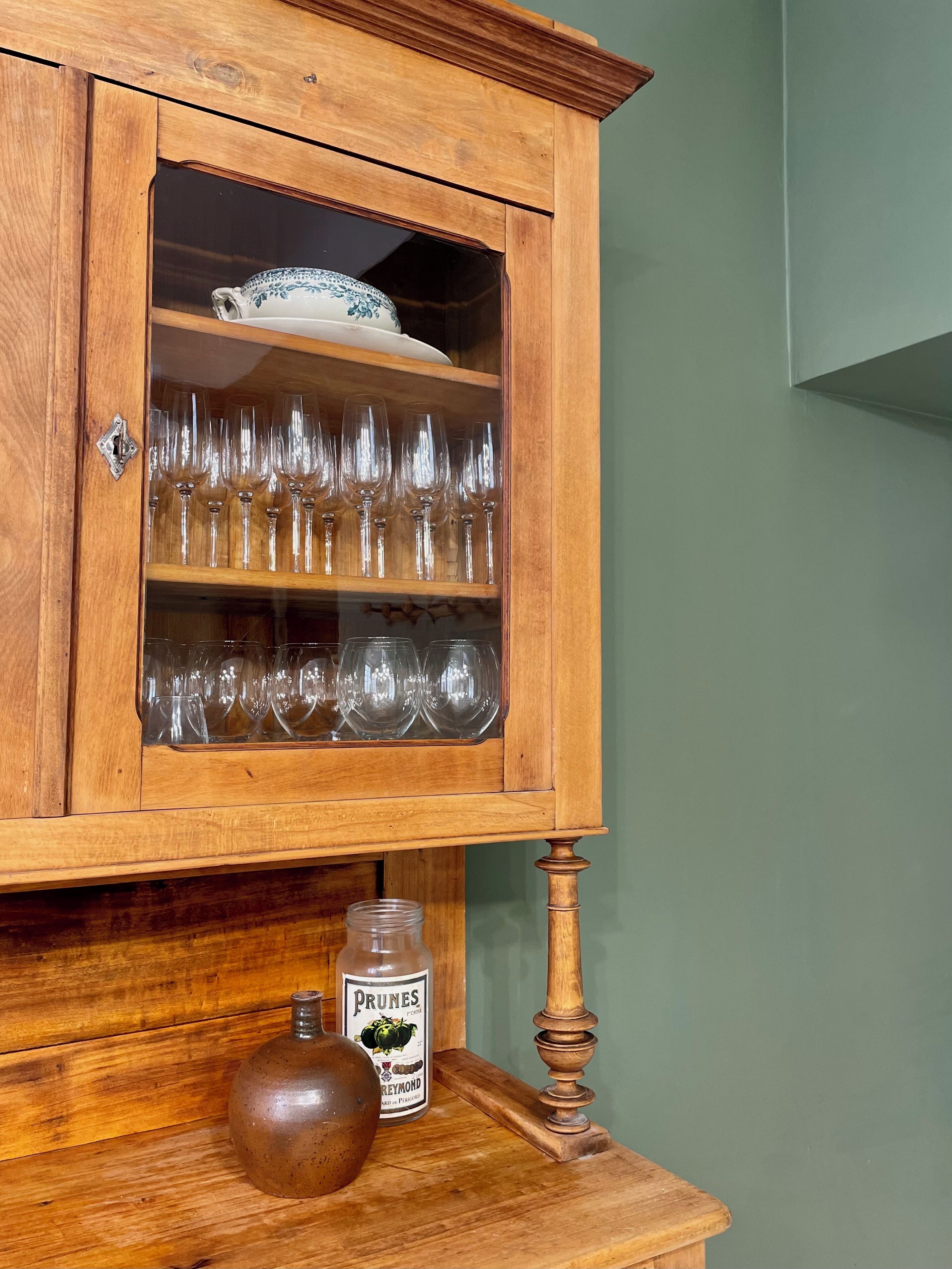 A wooden cabinet with glass doors containing wine glasses and a bottle of Prunes.