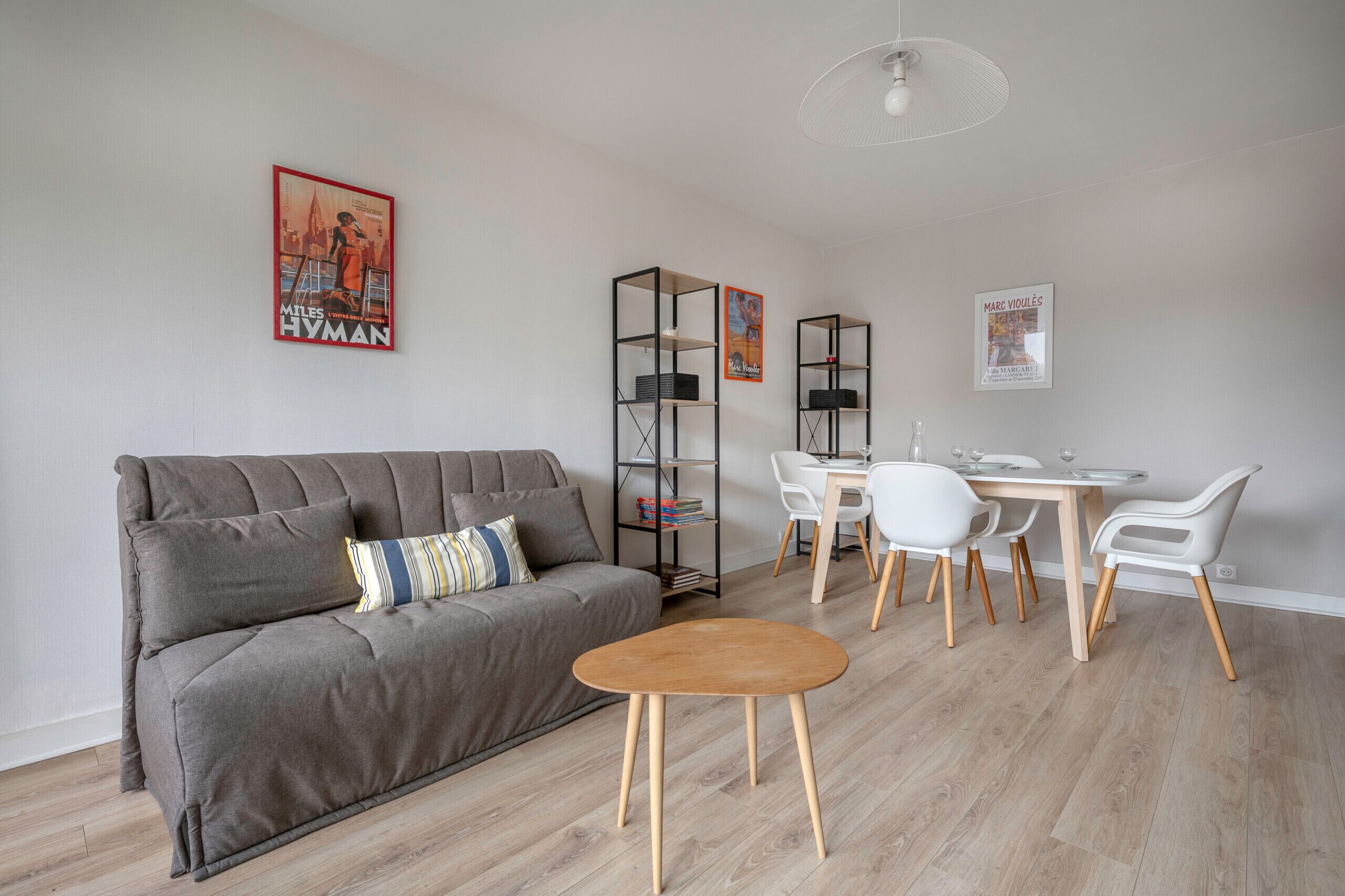 Ett modernt vardagsrum med en grå soffa, ett träbord och en matplats med vita stolar och ett träbord. Längs väggen står två svarta hyllsystem, och en takfläkt syns.