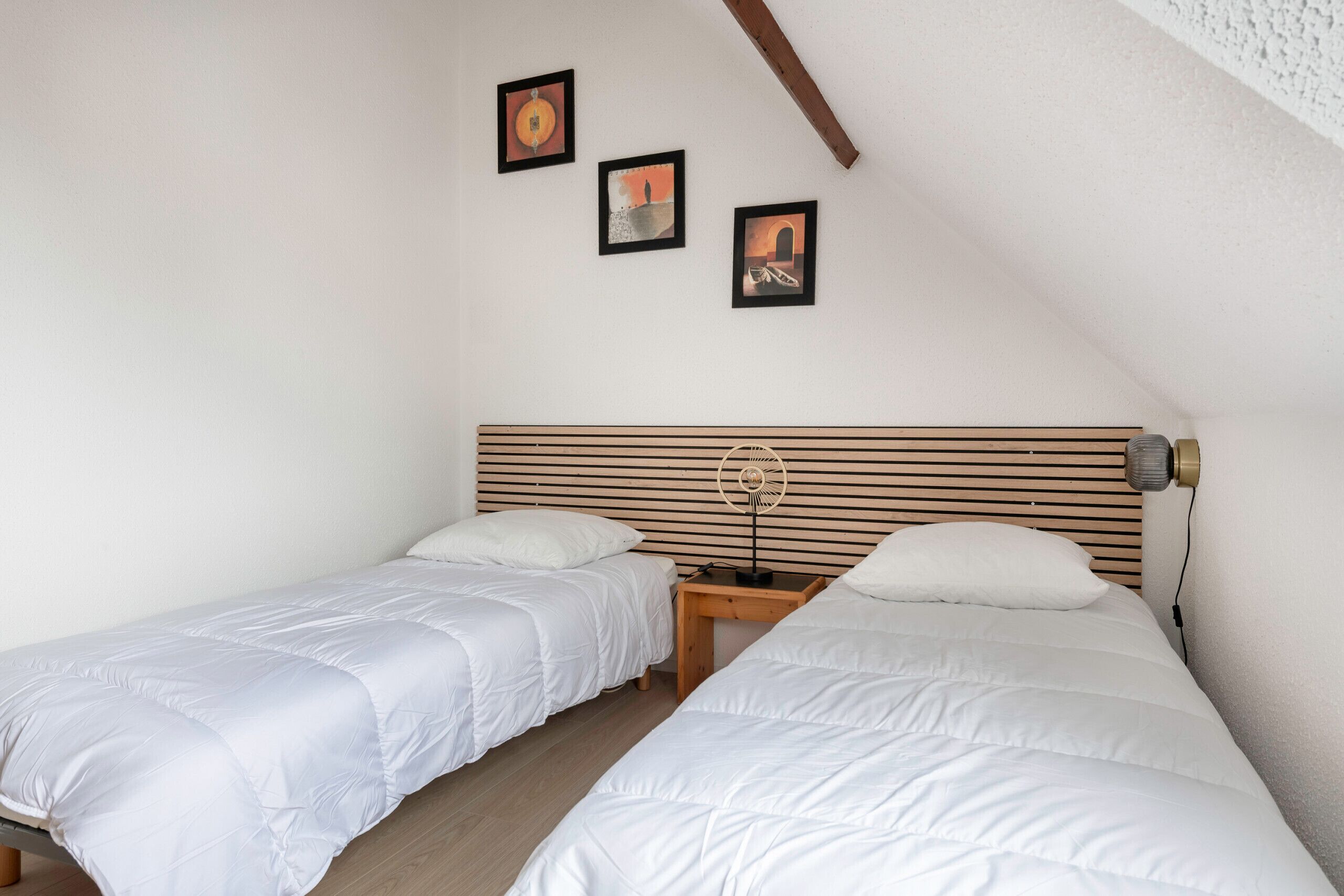 Une chambre avec deux lits simples, une tête de lit en bois, une table de chevet avec une lampe et trois tableaux encadrés accrochés au mur.