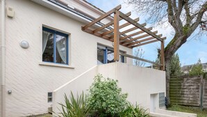 Una casa moderna con fachada blanca, una pérgola de madera y un pequeño jardín con diversas plantas.