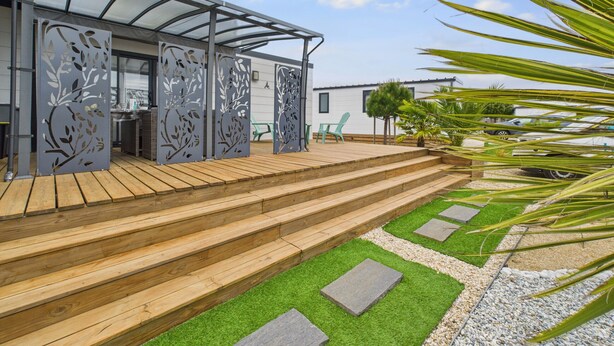 A modern outdoor patio with wooden steps leading to a covered seating area, surrounded by greenery and a clear sky. - Pretty Chalet for 6- Estate with swimming pool (Bretignolles-sur-Mer)