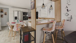 A modern kitchen with white cabinets, a wooden island, and pendant lights. There is a dining area with patterned chairs and a table. - Pretty Chalet for 6- Estate with swimming pool (Bretignolles-sur-Mer)
