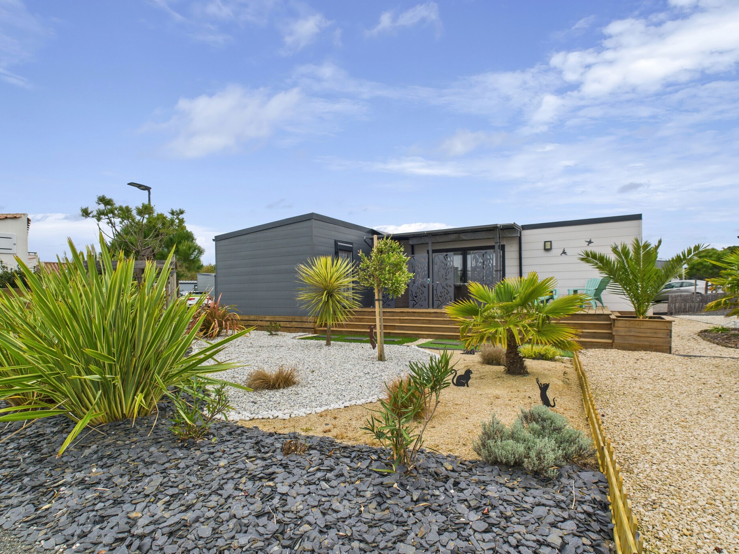 Une maison moderne à l’extérieur gris, dotée d’une terrasse en bois et d’un jardin de gravier agrémenté de différentes plantes.