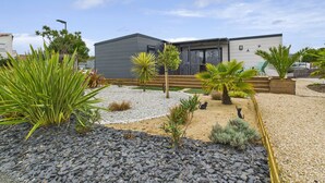 A modern house with a grey exterior, a wooden deck, and a gravel garden with various plants. - Pretty Chalet for 6- Estate with swimming pool (Bretignolles-sur-Mer)