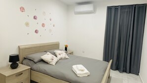A neatly made bed with a grey mattress, wooden headboard, and two pillows. There is a bedside table with a lamp and a small vase with flowers. The room has white walls, a tiled floor, and a window with grey curtains. - Central Cozy Located Apt (Msida)