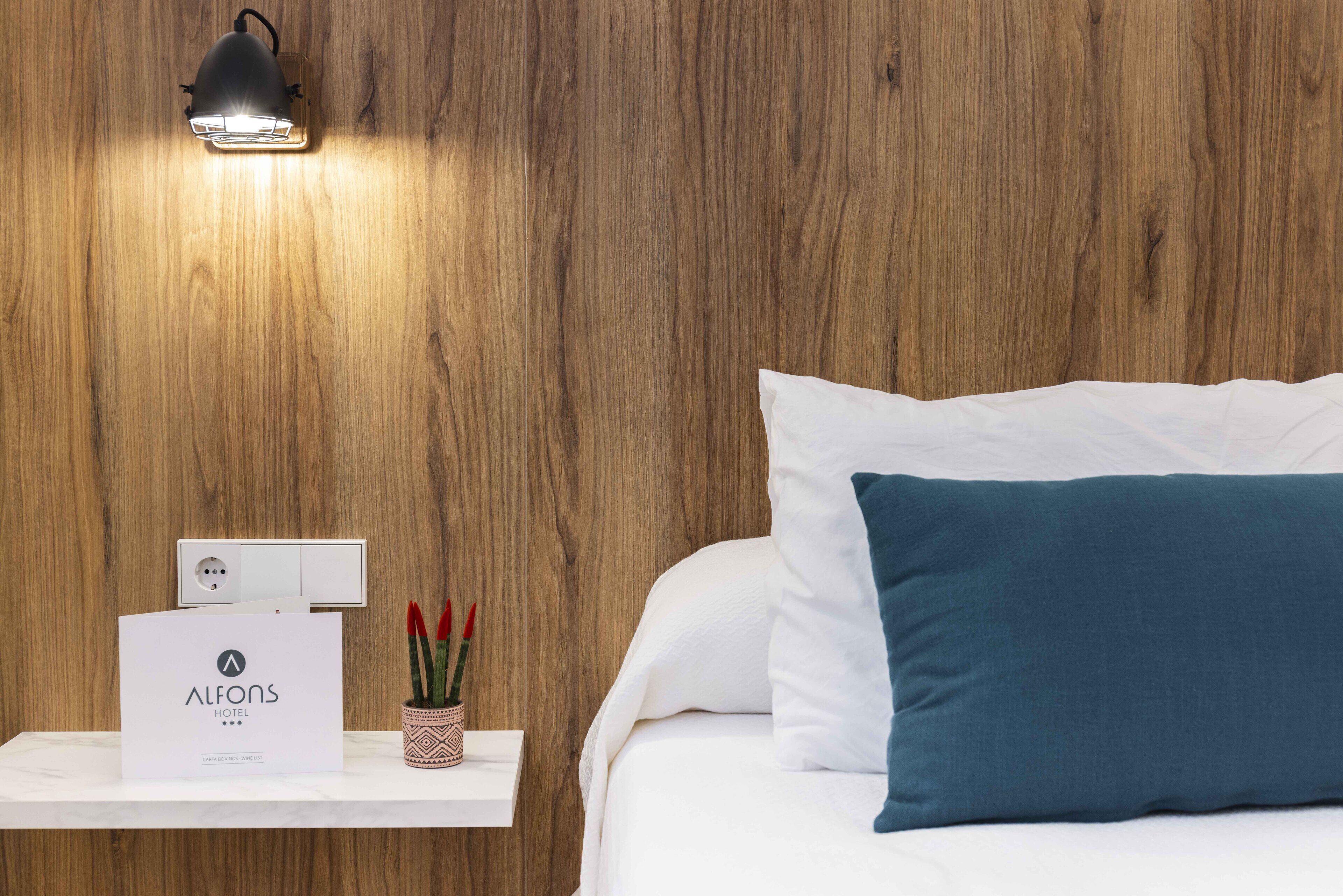 Une chambre d’hôtel avec un mur en bois, une lampe de chevet, une table de chevet blanche avec un panneau, un oreiller turquoise et un lit blanc.
