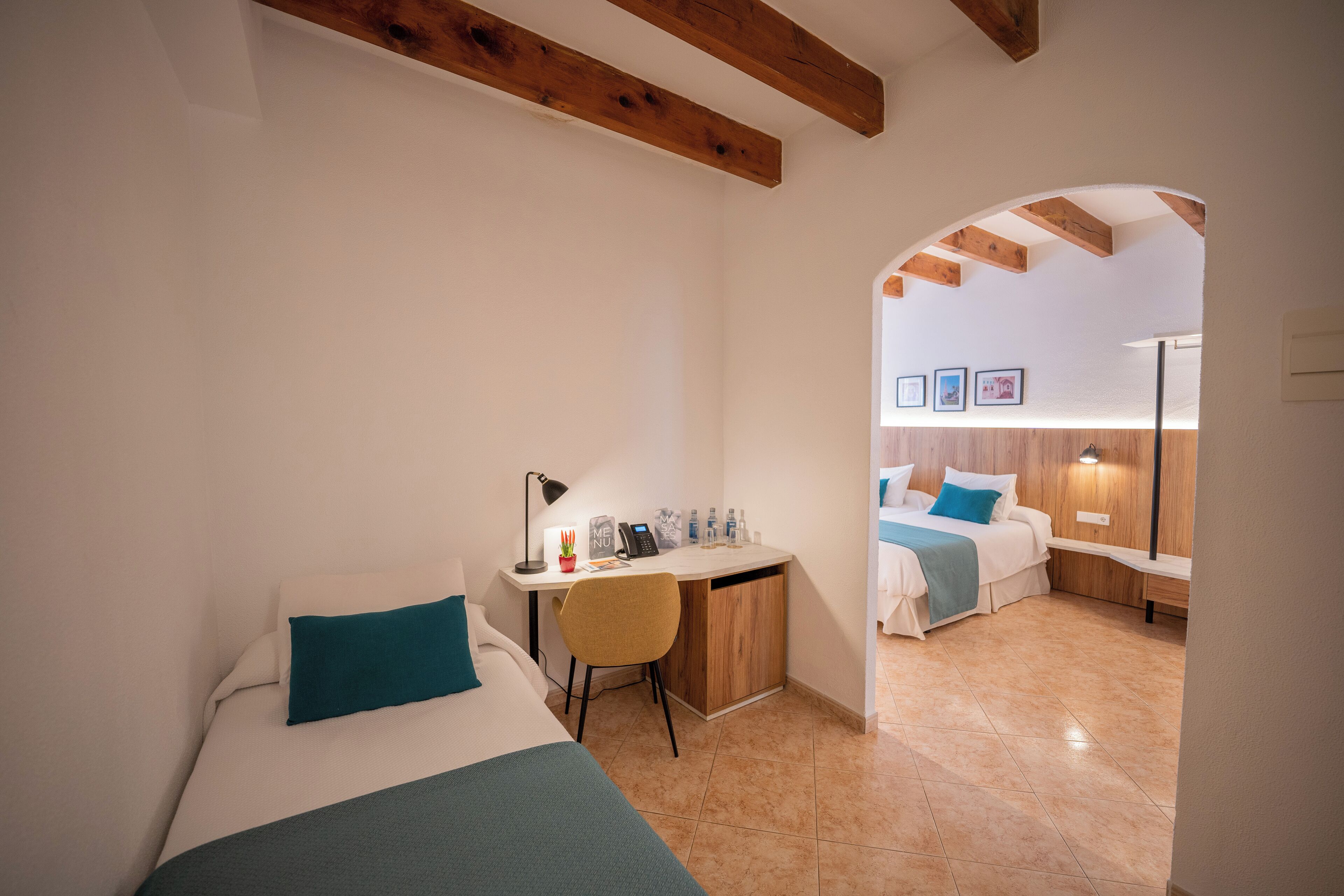 A compact hotel room with a single bed, a desk, and a chair. There is a small table with a lamp and a few items on it. The room has wooden beams on the ceiling and a tiled floor.