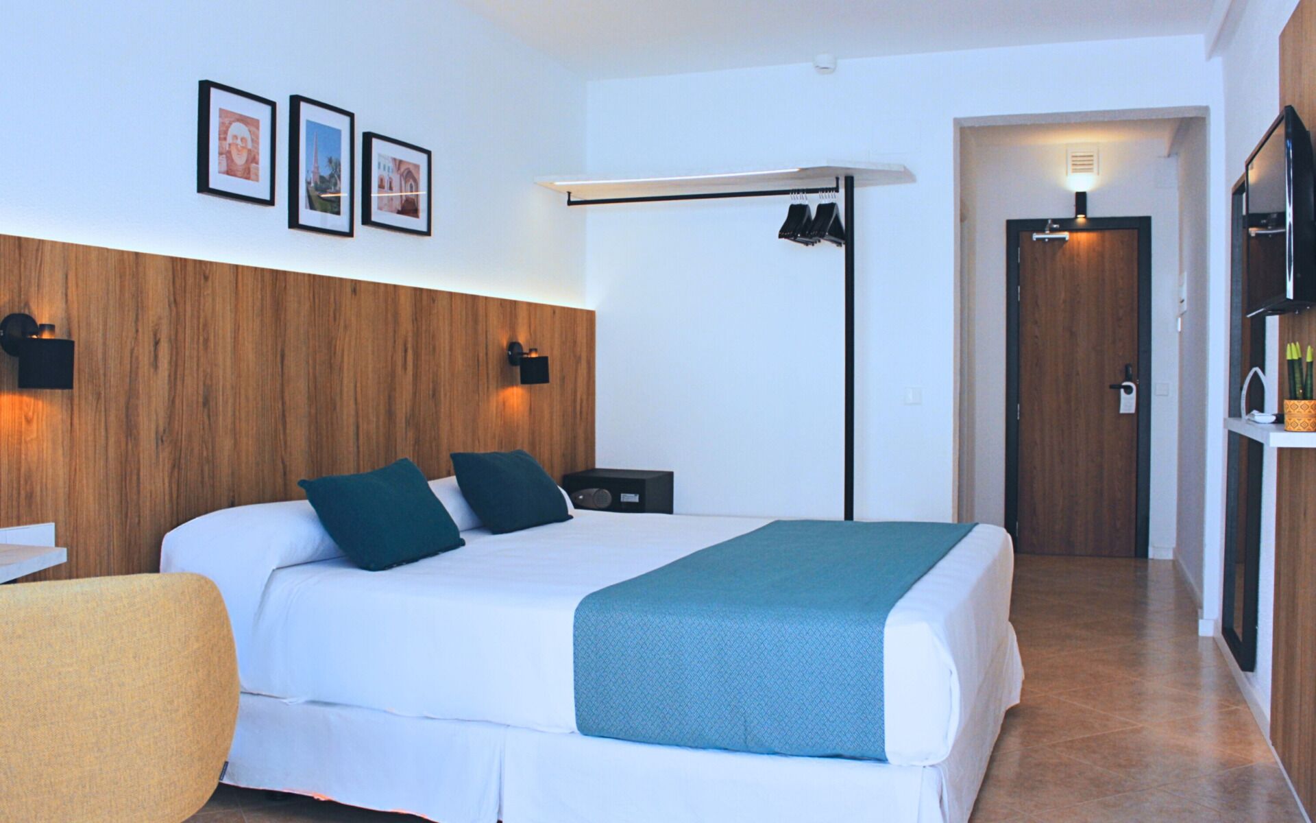 Habitación de hotel con una cama grande, un escritorio, una silla, un armario y cuadros en la pared.