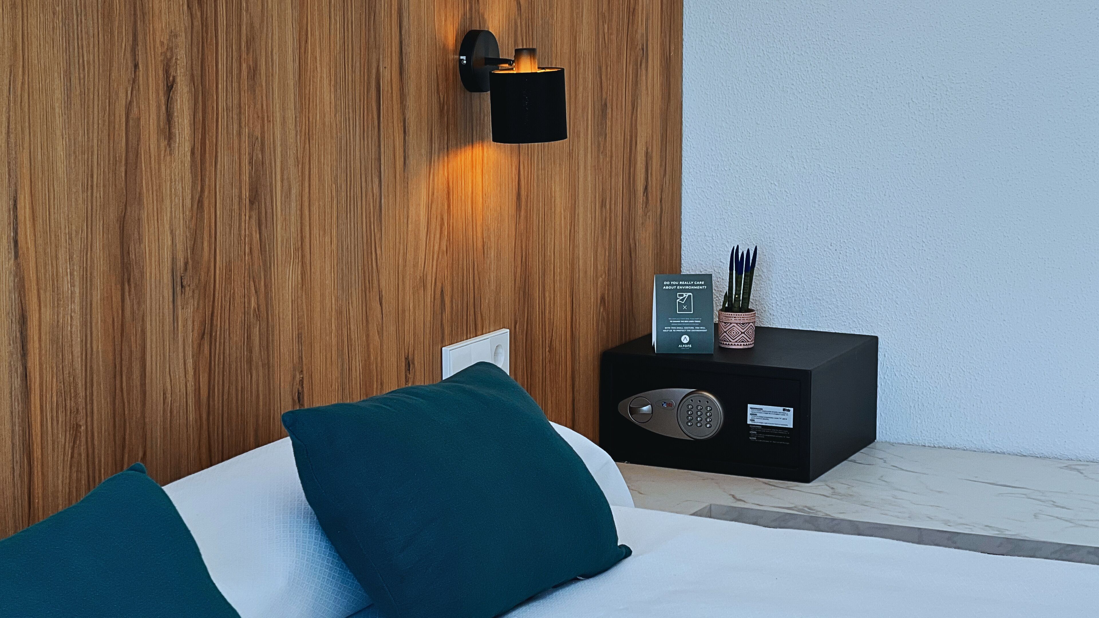 Ein modernes Zimmer mit einer Wand in Holzoptik, einem schwarzen Nachttisch mit Radio und türkisfarbenen Kissen auf einem weißen Bett.