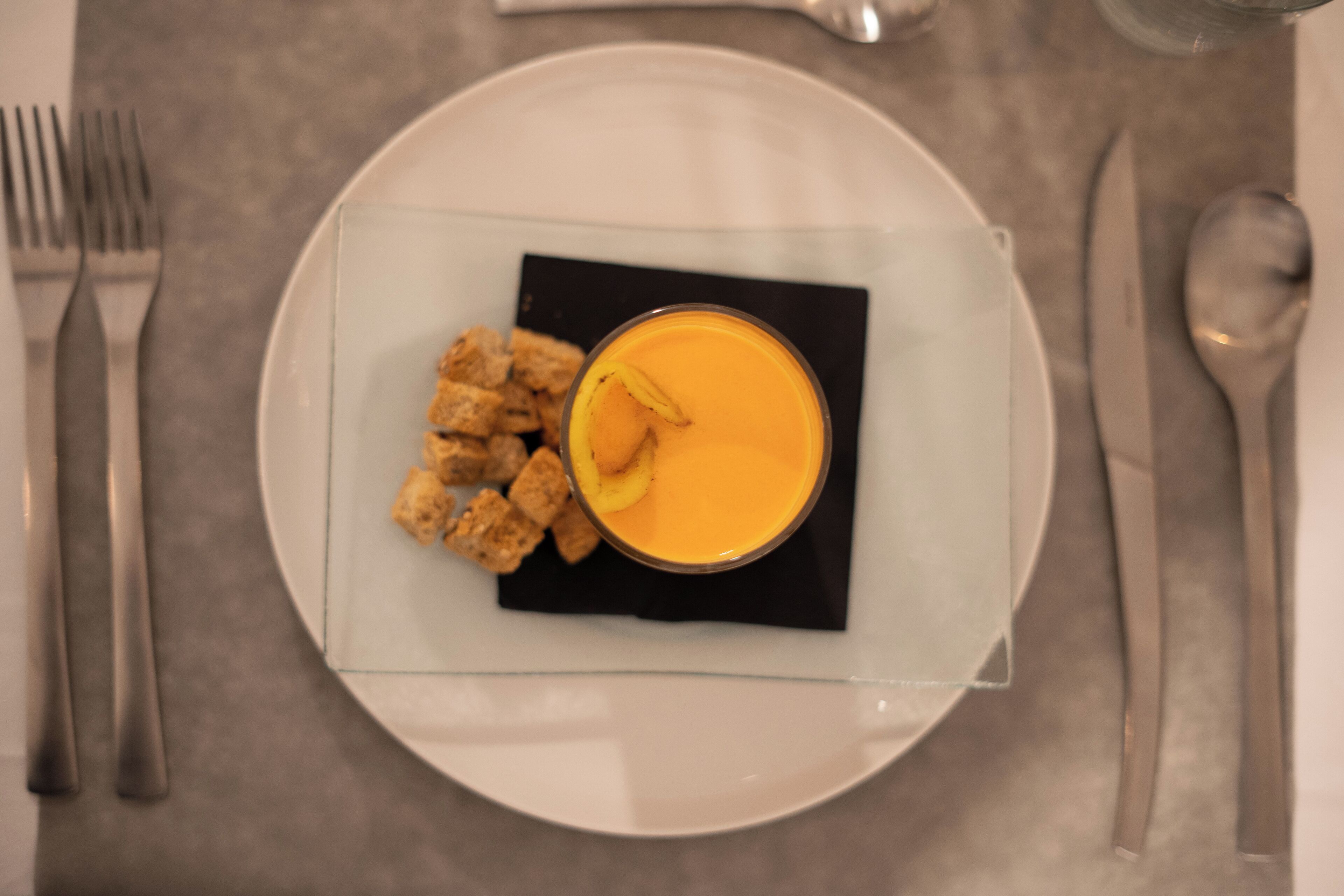 Un plato con un vaso de líquido naranja, posiblemente un batido o jugo, y algunos croutons sobre un plato de vidrio transparente.