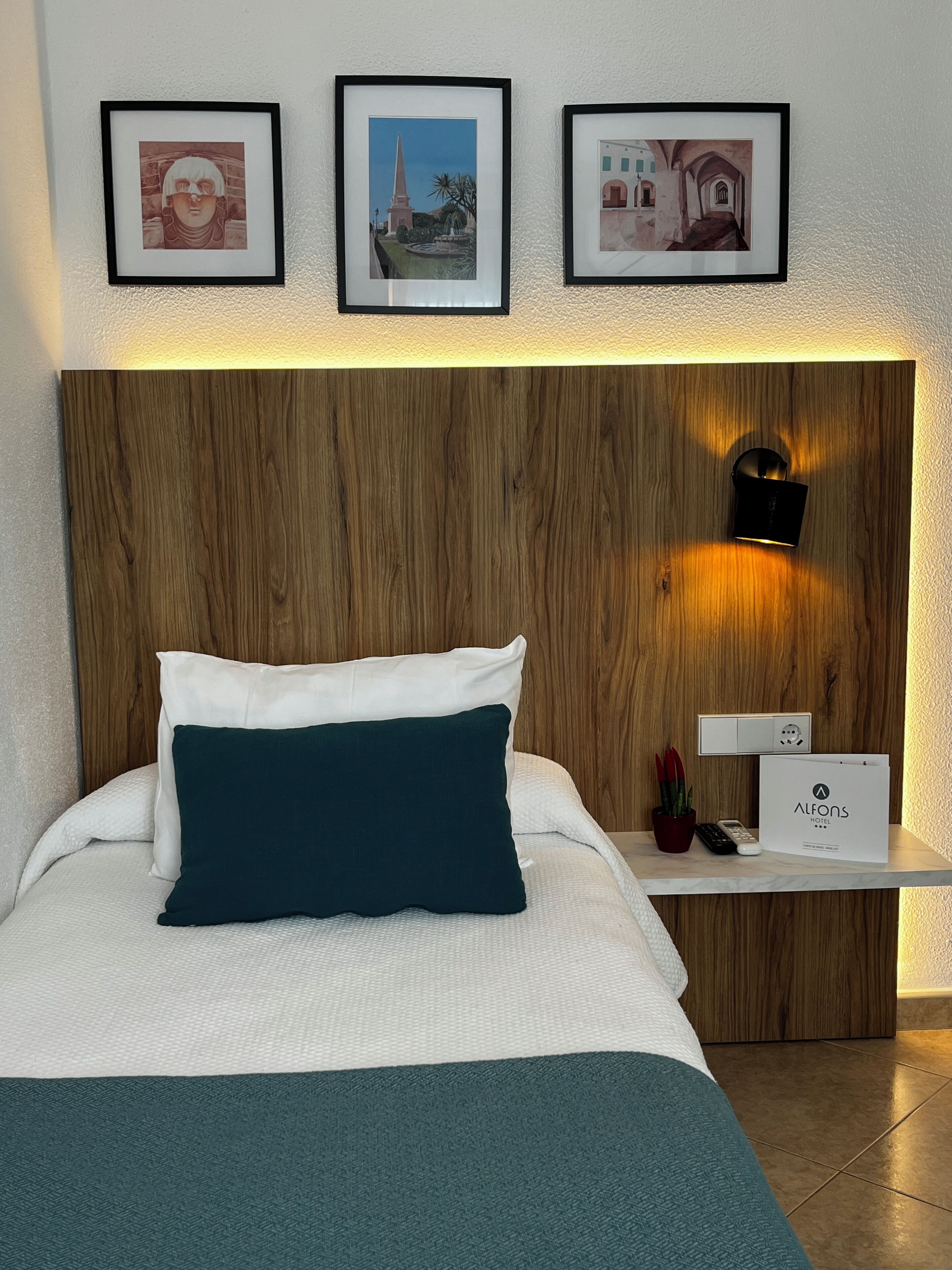 Camera d'albergo con una testiera in legno, un letto con biancheria da letto bianco e verde scuro, un comodino con un telecomando, una pianta e un quadro incorniciato sopra il letto.