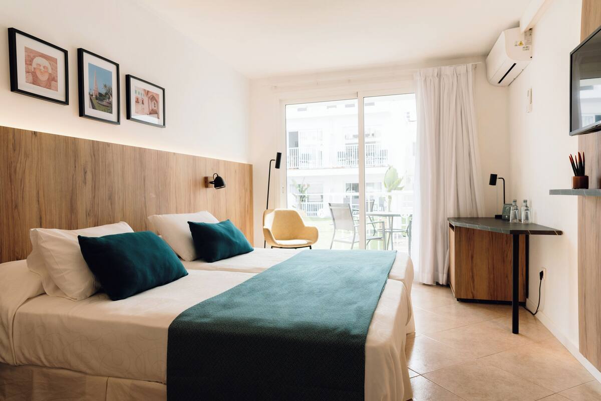Une chambre d’hôtel avec deux lits, un bureau, une chaise et un balcon donnant sur des immeubles.