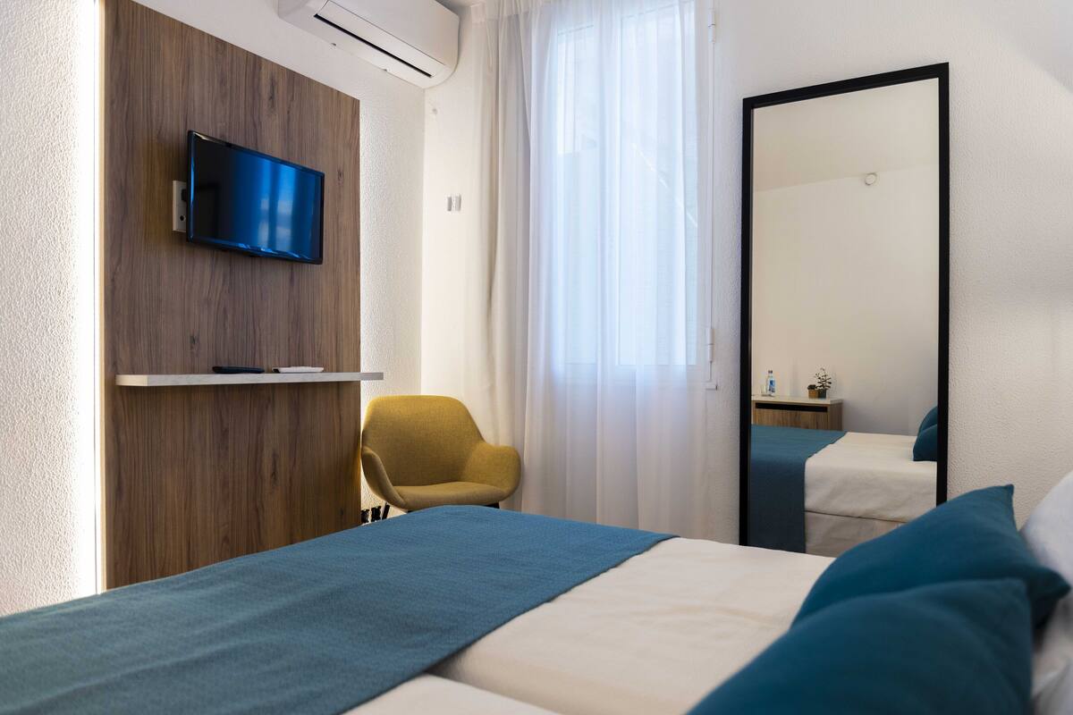 Une chambre d’hôtel avec un lit, un téléviseur fixé sur un panneau en bois, un fauteuil jaune, un grand miroir et une fenêtre avec des rideaux légers.