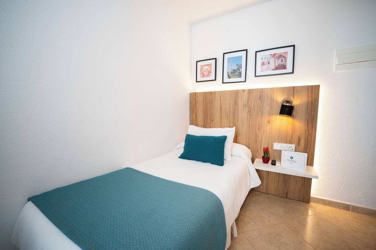 Une chambre d’hôtel bien rangée, avec un lit, une table de chevet et une étagère fixée au mur, sur laquelle est posé un panneau.