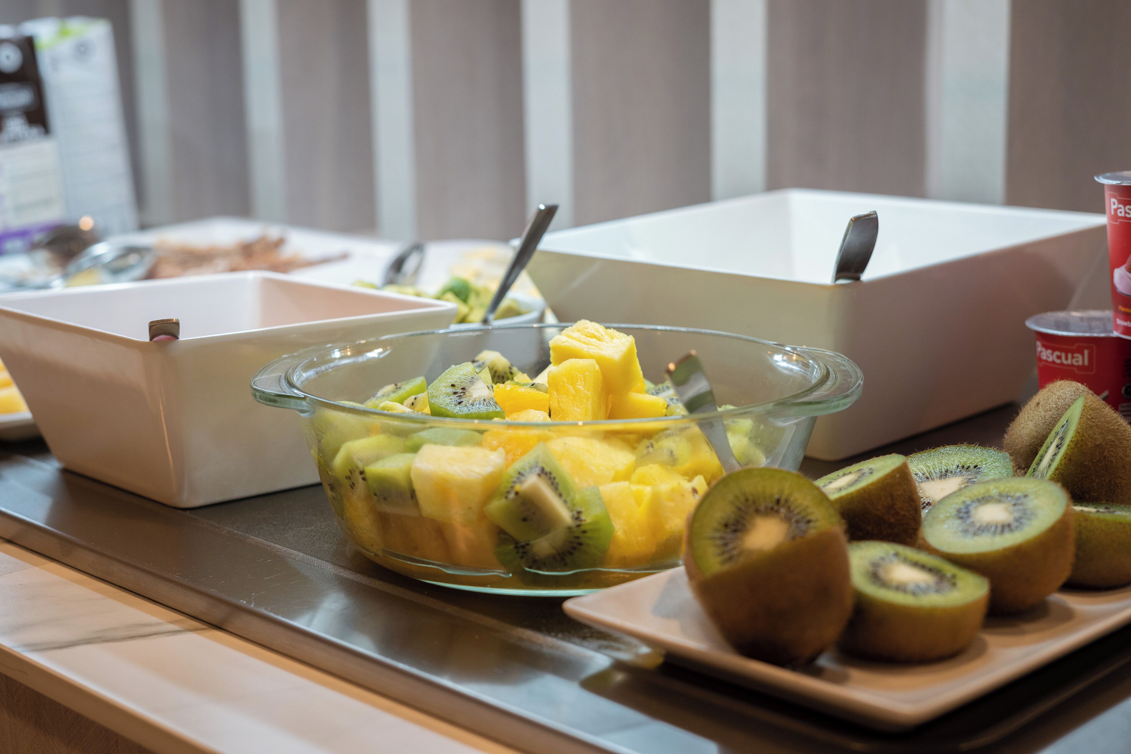Una mesa de buffet con un bol de vidrio lleno de frutas cortadas y un plato con rodajas de kiwi.