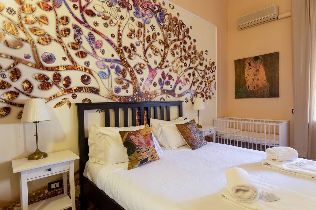 A hotel room with a large decorative wall, a crib, a neatly made bed with white linens, and a painting on the wall.