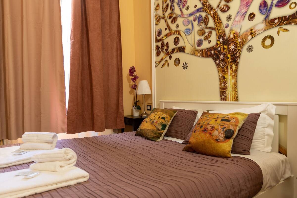 A neatly made bed with a patterned bedspread, pillows with a floral design, and a wall tapestry depicting a tree.