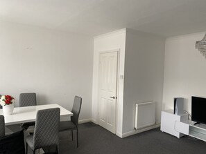 Habitación moderna con una mesa blanca, sillas grises, una puerta blanca, un radiador y un mueble para televisión.