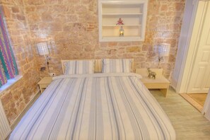 A bedroom with a stone wall, a bed with striped bedding, a nightstand with a lamp, and a shelf with a decorative item. -  Deniz Önü Guesthouse (Ayvalik)