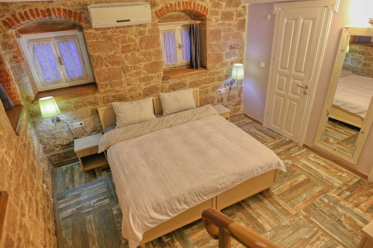 A bedroom with a stone wall, a wooden bed, a window with curtains, a bedside lamp, and a door.