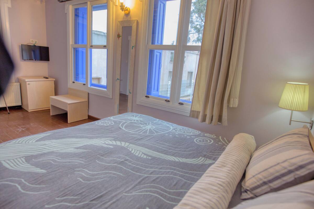 A bedroom with a bed, a bedside table, a lamp, a TV, and a window with curtains.