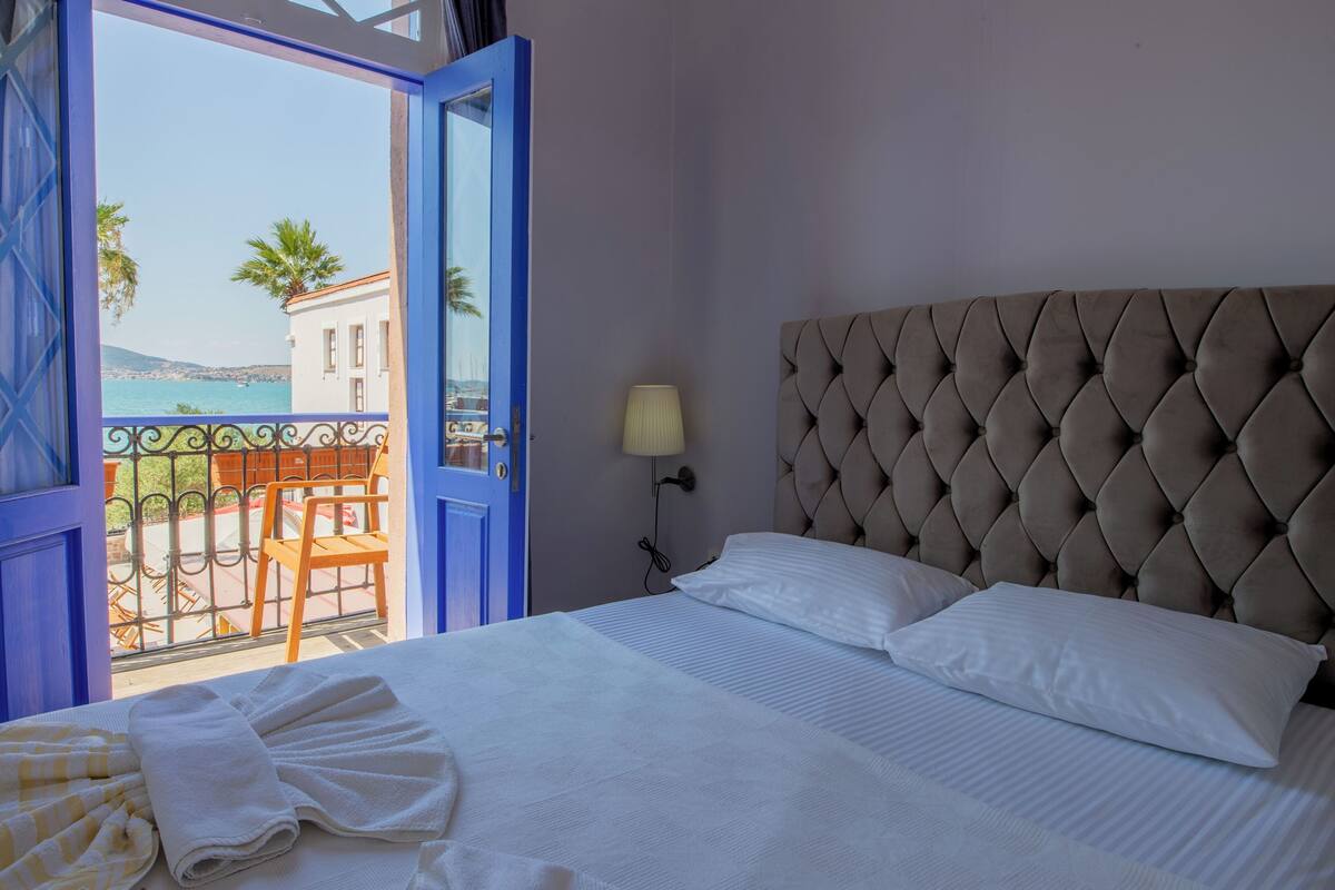 A hotel room with a bed, a view of the sea, and a balcony with chairs.