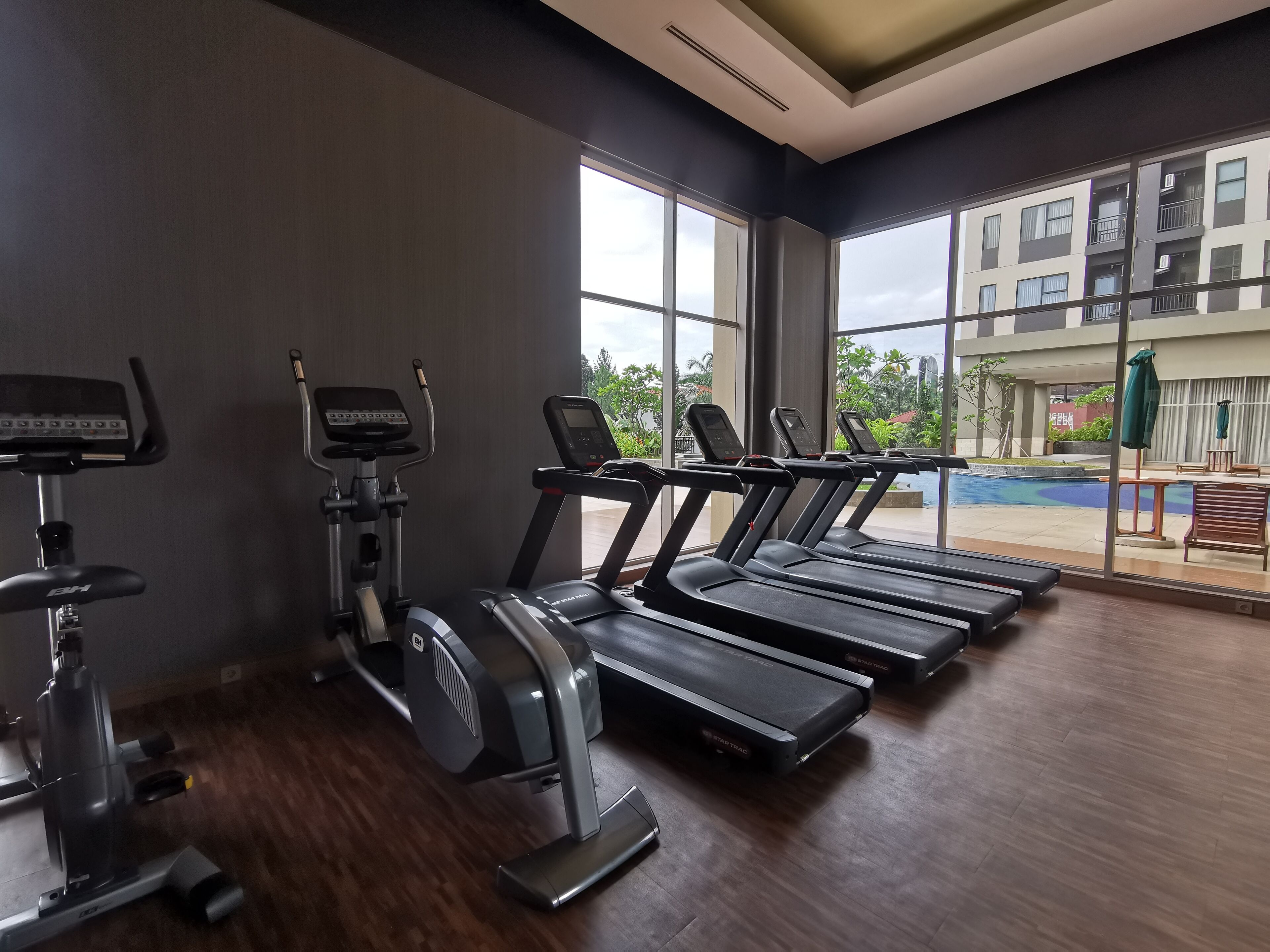 A gym with treadmills, an elliptical, and a stationary bike. Large windows offer a view of a building and greenery outside.