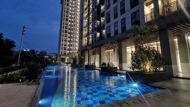 A swimming pool with a tiled deck, surrounded by buildings with balconies and outdoor lighting.