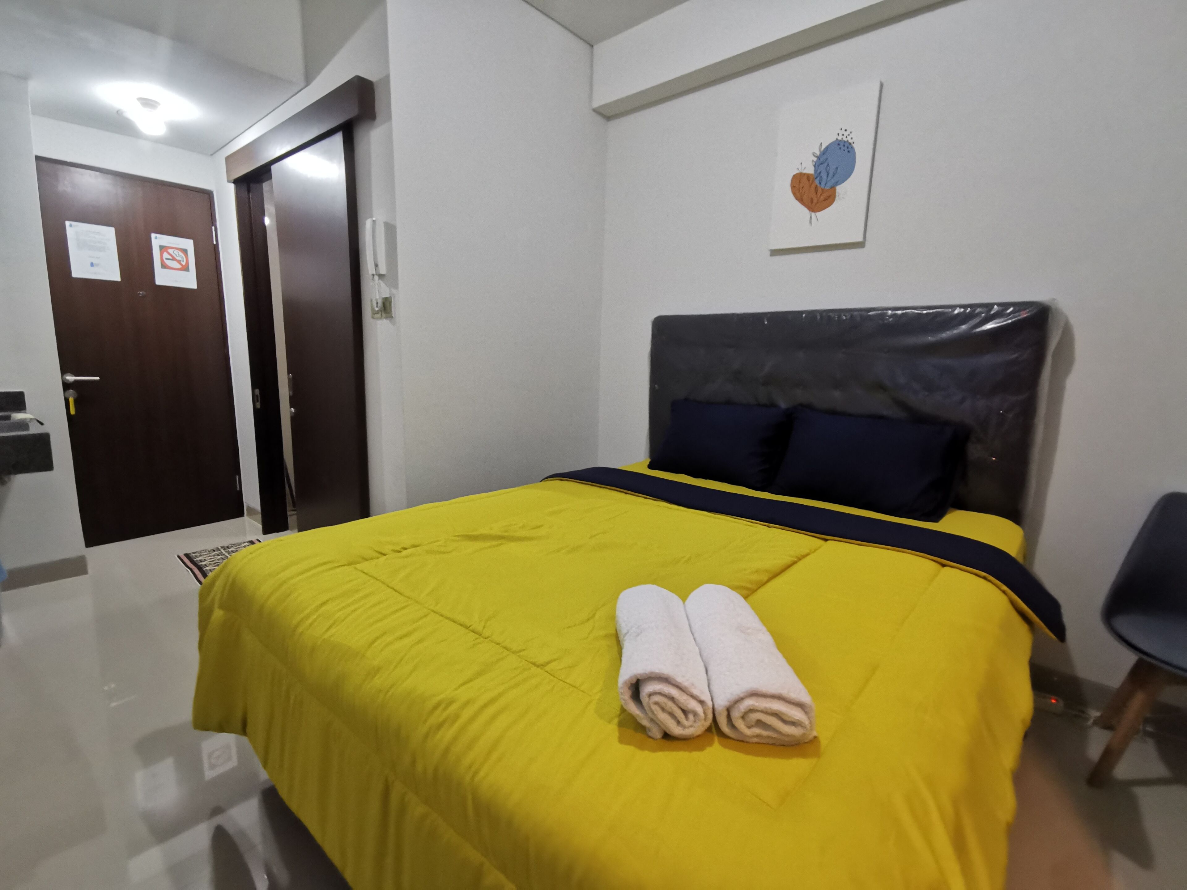 Habitación de hotel con colcha amarilla, dos toallas enrolladas, cabecero oscuro y una obra de arte colgada en la pared.