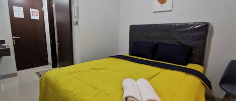 Habitación de hotel con colcha amarilla, dos toallas enrolladas, cabecero oscuro y una obra de arte colgada en la pared.
