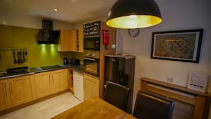 A compact kitchen with wooden cabinets, a dining table, a refrigerator, and a hanging light fixture.