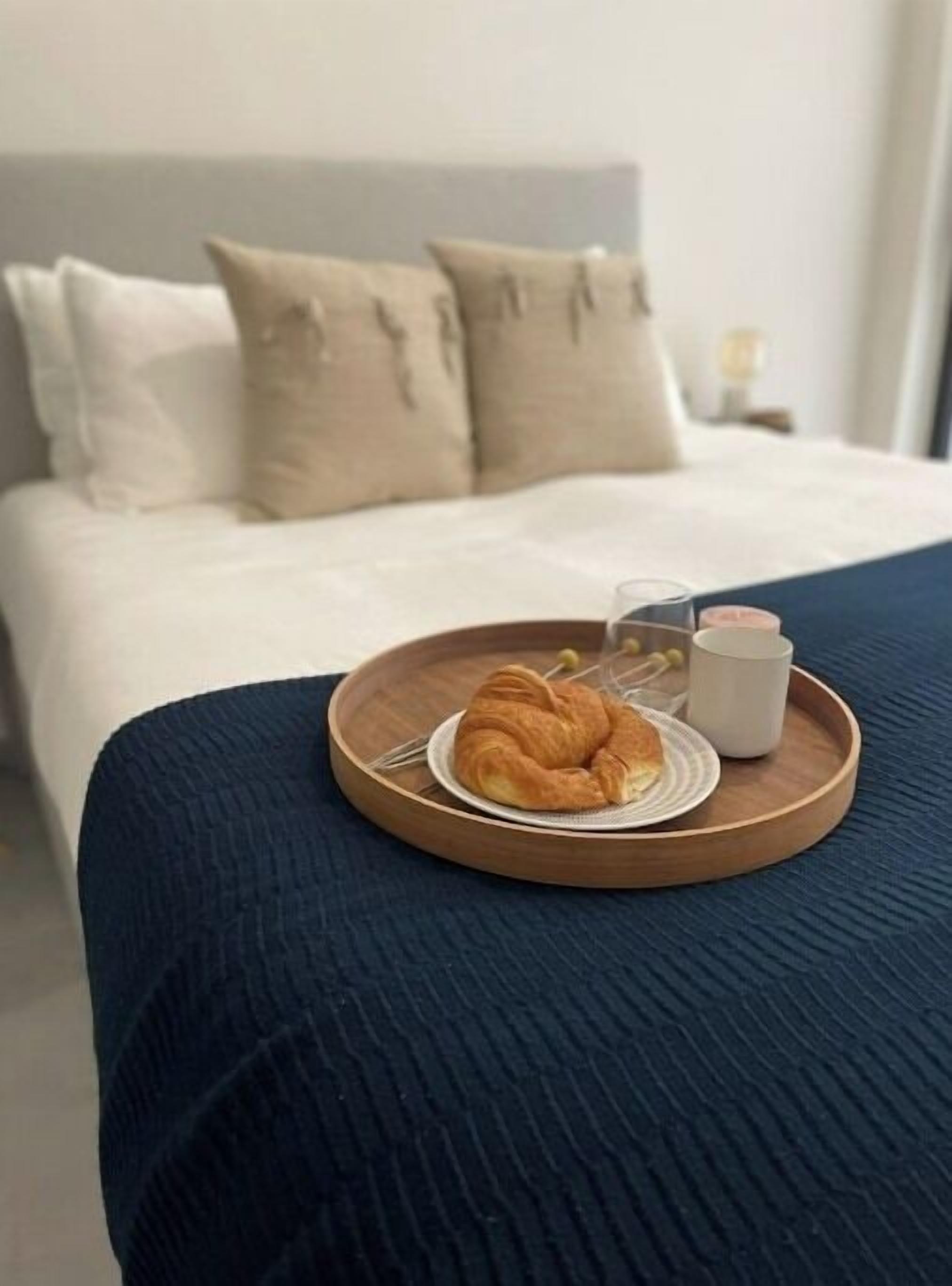 A bed with a dark blue bedspread, a wooden tray with a croissant, a glass, and a white mug on it.