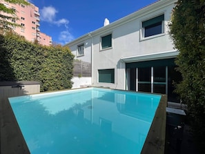 A swimming pool with a clear blue water surface, surrounded by a hedge and a building with a garage door.
