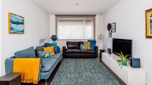 A modern living room with a black leather sofa, blue and orange cushions, a patterned rug, a television, and a potted plant. - Clementina: Serene Garden and Pool Stay w/Parking (Lisboa)