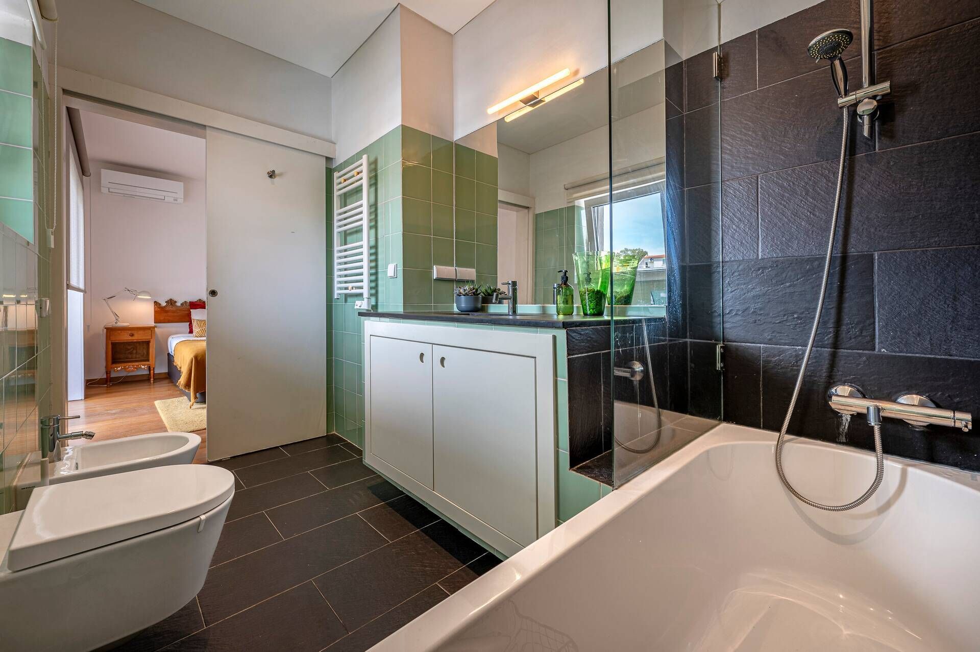 A modern bathroom with a bathtub, a shower, and a toilet. There is a vanity with a mirror and a countertop. The walls are tiled in two colors, and there is a window providing natural light.