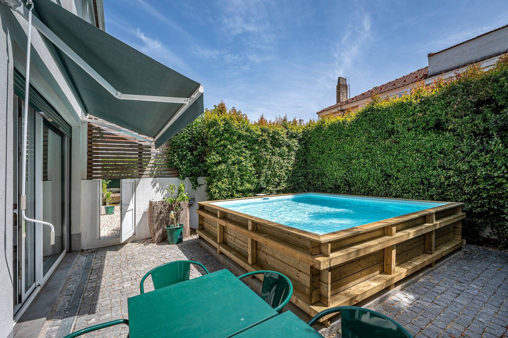 A wooden hot tub in a backyard with a brick patio, a wooden deck, and a green table and chairs.