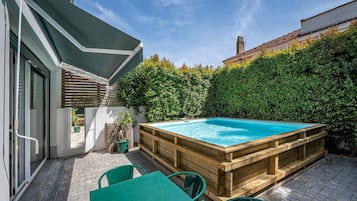 A wooden hot tub in a backyard with a brick patio, a wooden deck, and a green table and chairs.