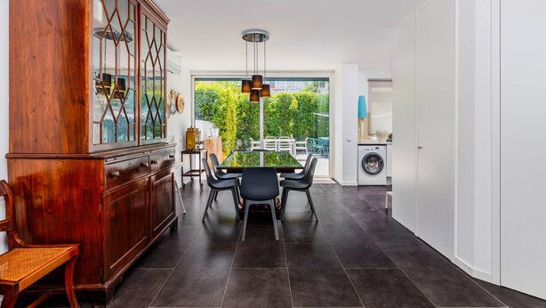 A modern kitchen with dark tiled flooring, a wooden cabinet with glass doors, a dining table with chairs, and a view of greenery outside. - Clementina: Serene Garden and Pool Stay w/Parking (Lisboa)