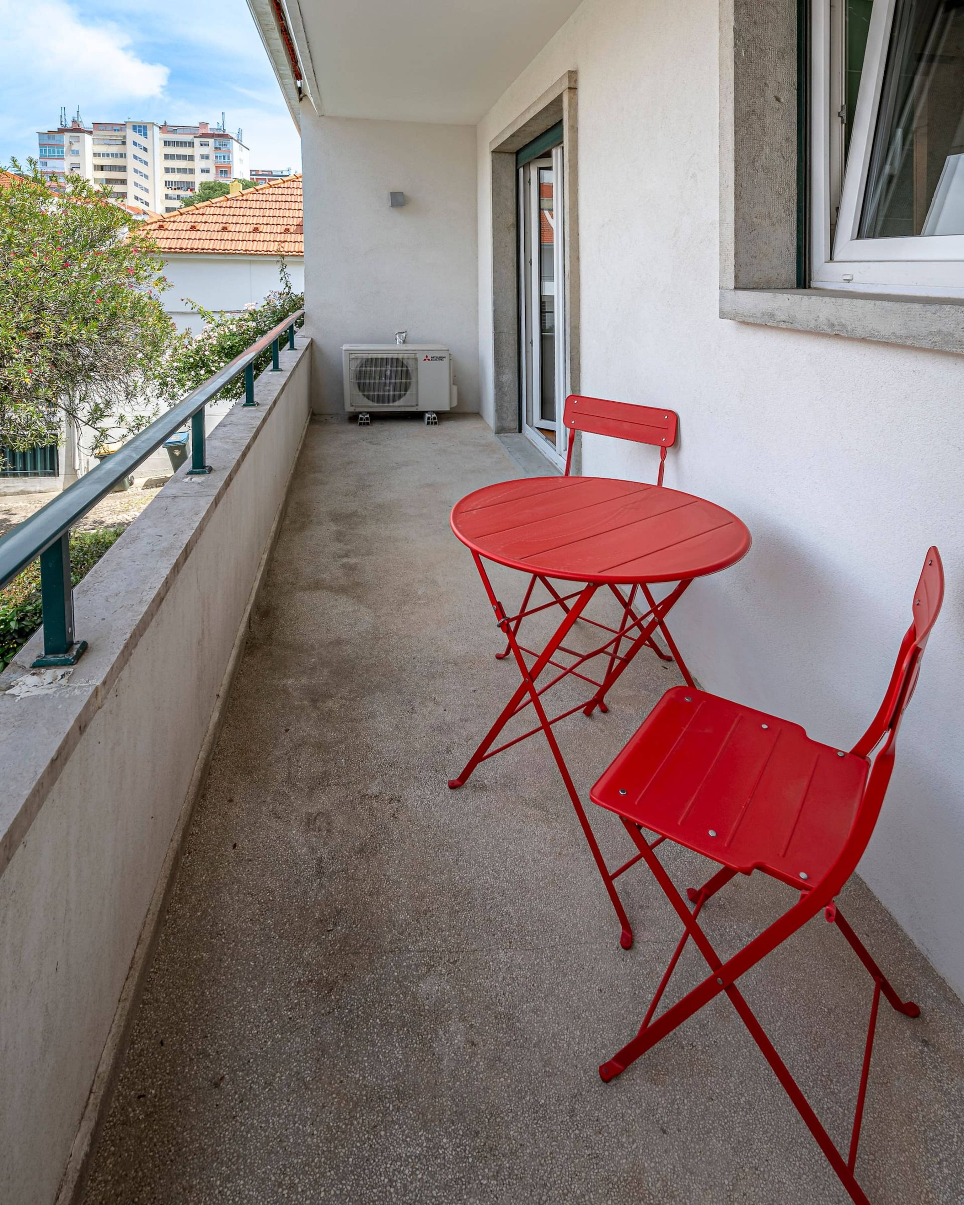 A balcony with a red metal table and chairs, an air conditioning unit, and a view of buildings in the background.