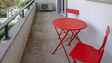 A balcony with a red metal table and chairs, an air conditioning unit, and a view of buildings in the background.