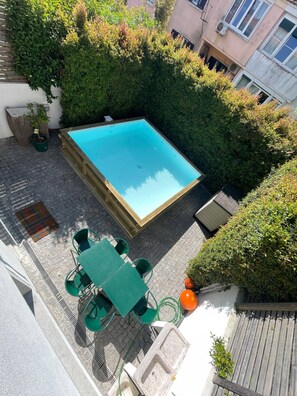 Una piscina en la azotea con terraza de madera, rodeada de abundante follaje y una pequeña mesa con sillas.