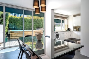 A modern kitchen with a built-in washing machine, a dining area with a glass table, and a view of a lush garden through large sliding doors.