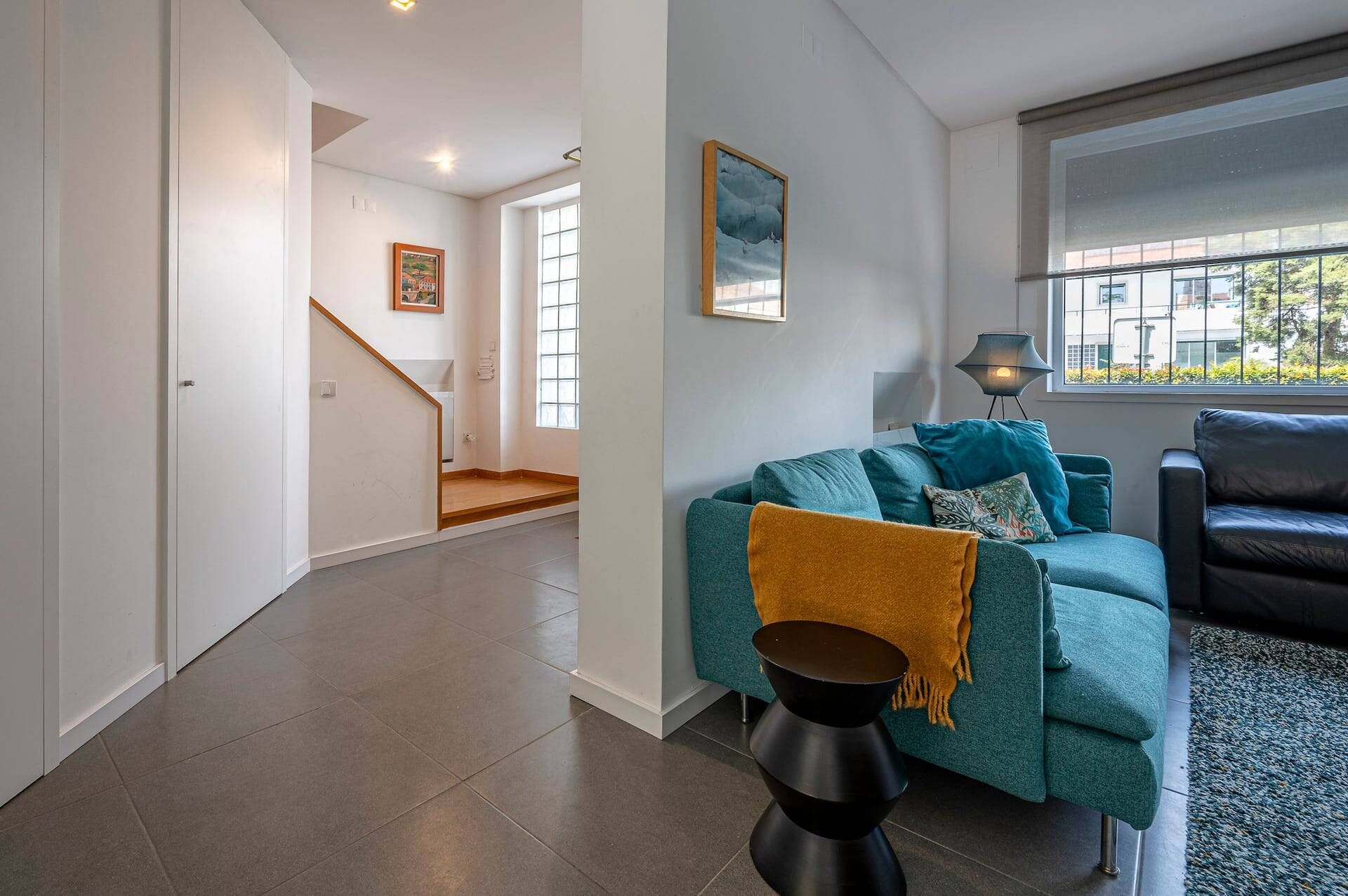 A modern living room with a teal sofa, a black coffee table, and a black leather couch. There is a staircase leading to another room, and a window with a view of the outside.